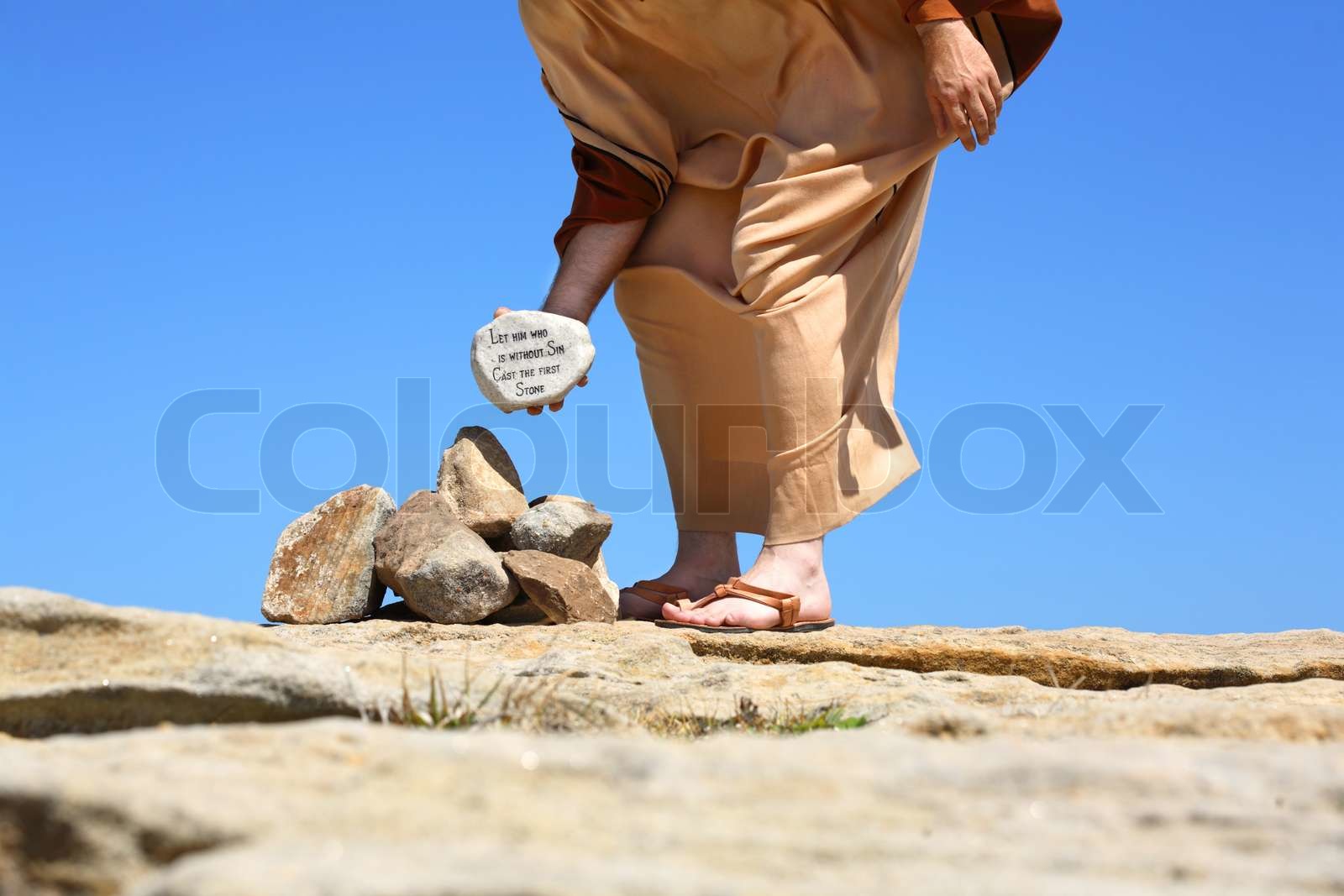 Man standing in the sun on sandy rocks holding a rock depicting ansswer - Hij Die Zonder Zonde Is Werpe De Eerste Steen